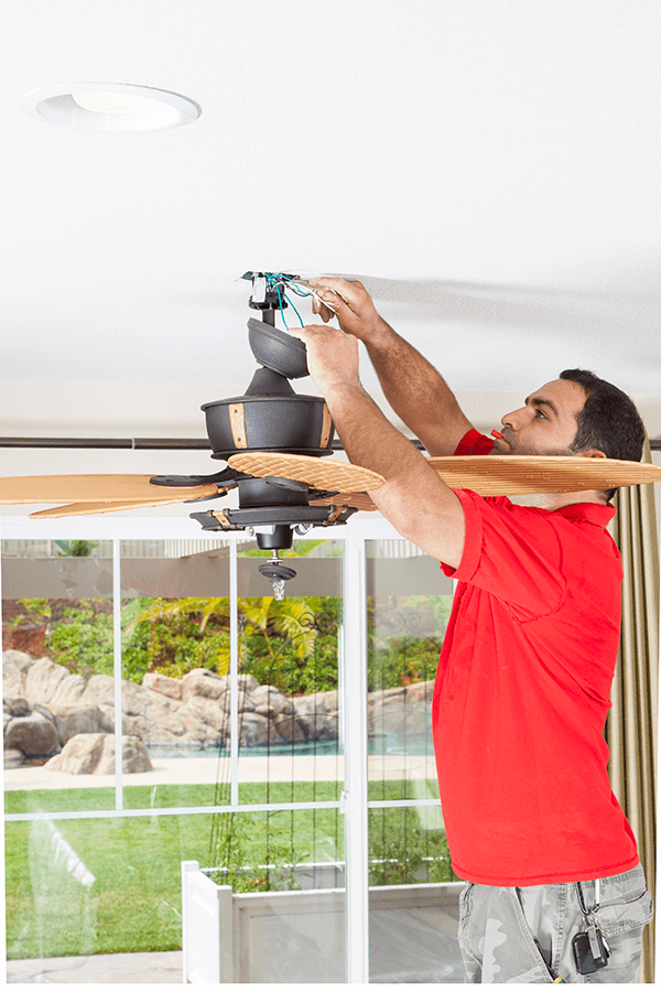 A man stands on a ladder, fixing a ceiling fan with focused concentration.