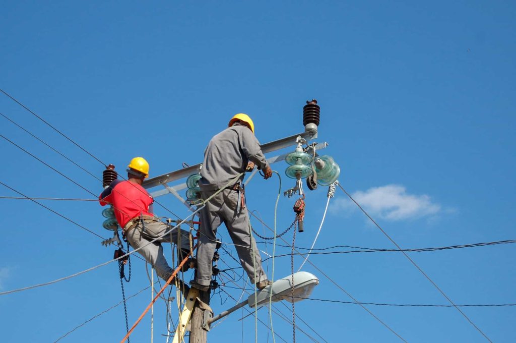 Two men wearing safety gear are working on an electrical pole, fixing wires and ensuring proper connections.